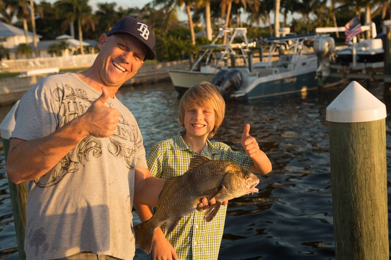 A man and a boy on a dock at golden hour holding a fish, boats and palms behind