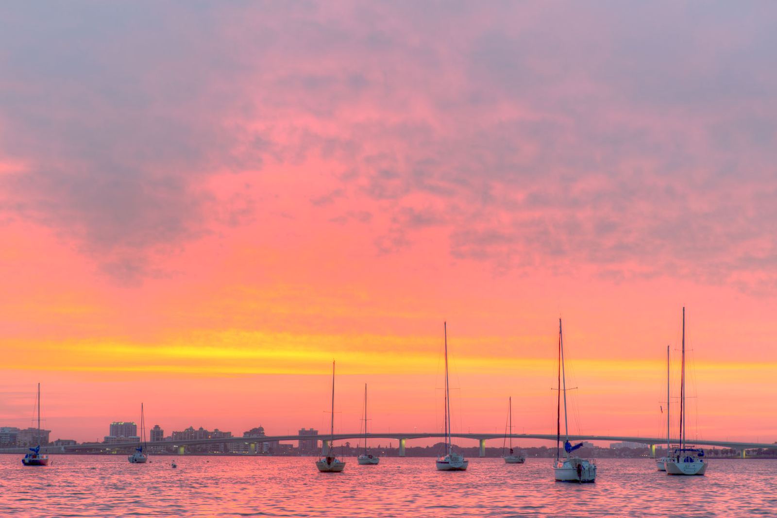 A pink and orange sunrise over Sarasota Bay with moored sailboats and the Ringling Causeway