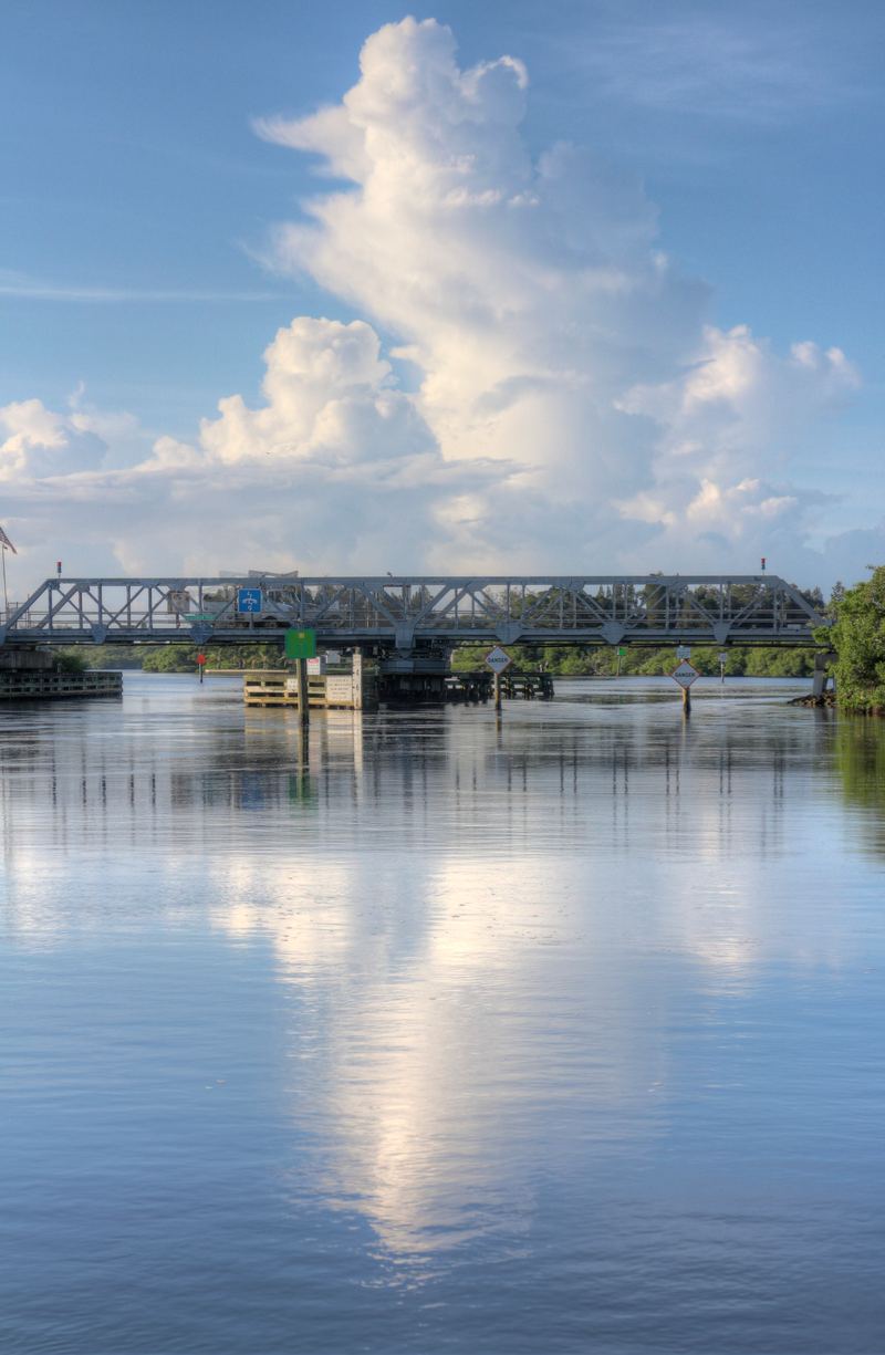 A one-lane steel truss swing bridge across a narrow waterway, with fender pilings below the span and a thunderhead building behind it