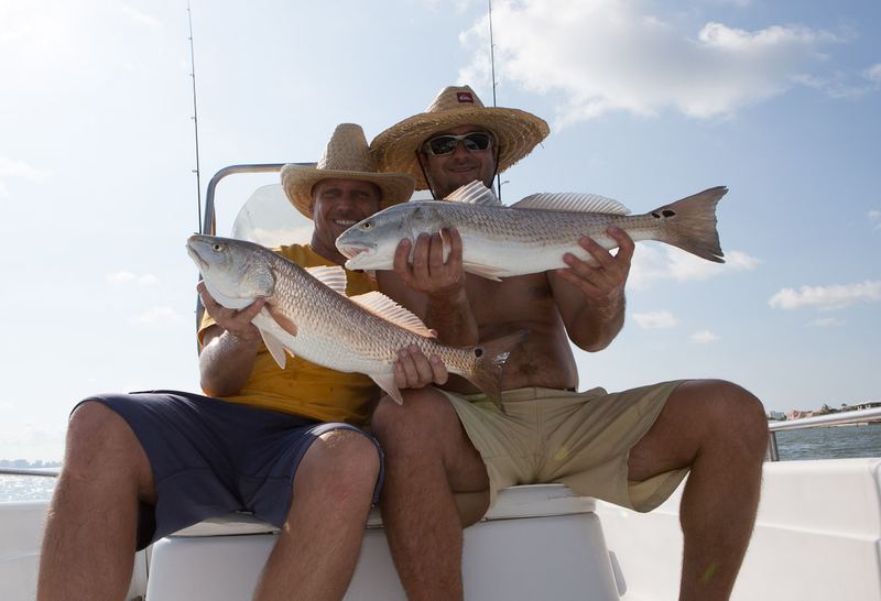 Two anglers seated on a boat gunwale, each holding a redfish