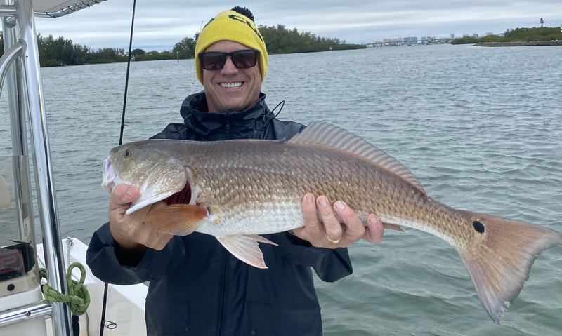 A big redfish held boat-side on a gray, cold-front morning, angler in a beanie and foul-weather jacket