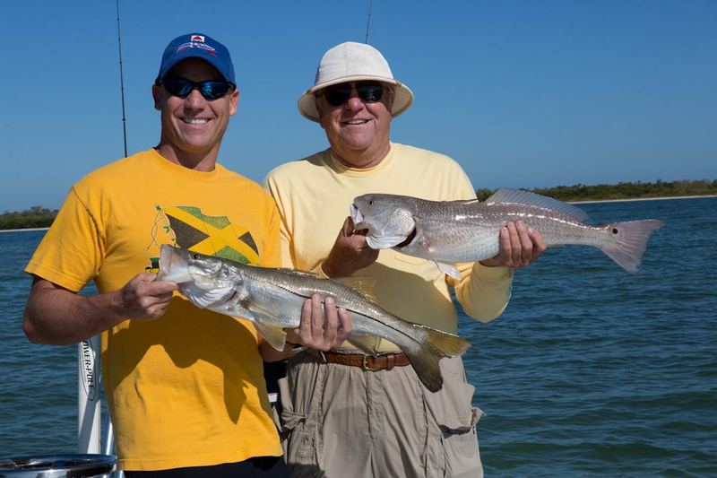 Two anglers on a boat, one holding a snook and the other a redfish, bay and mangrove island behind