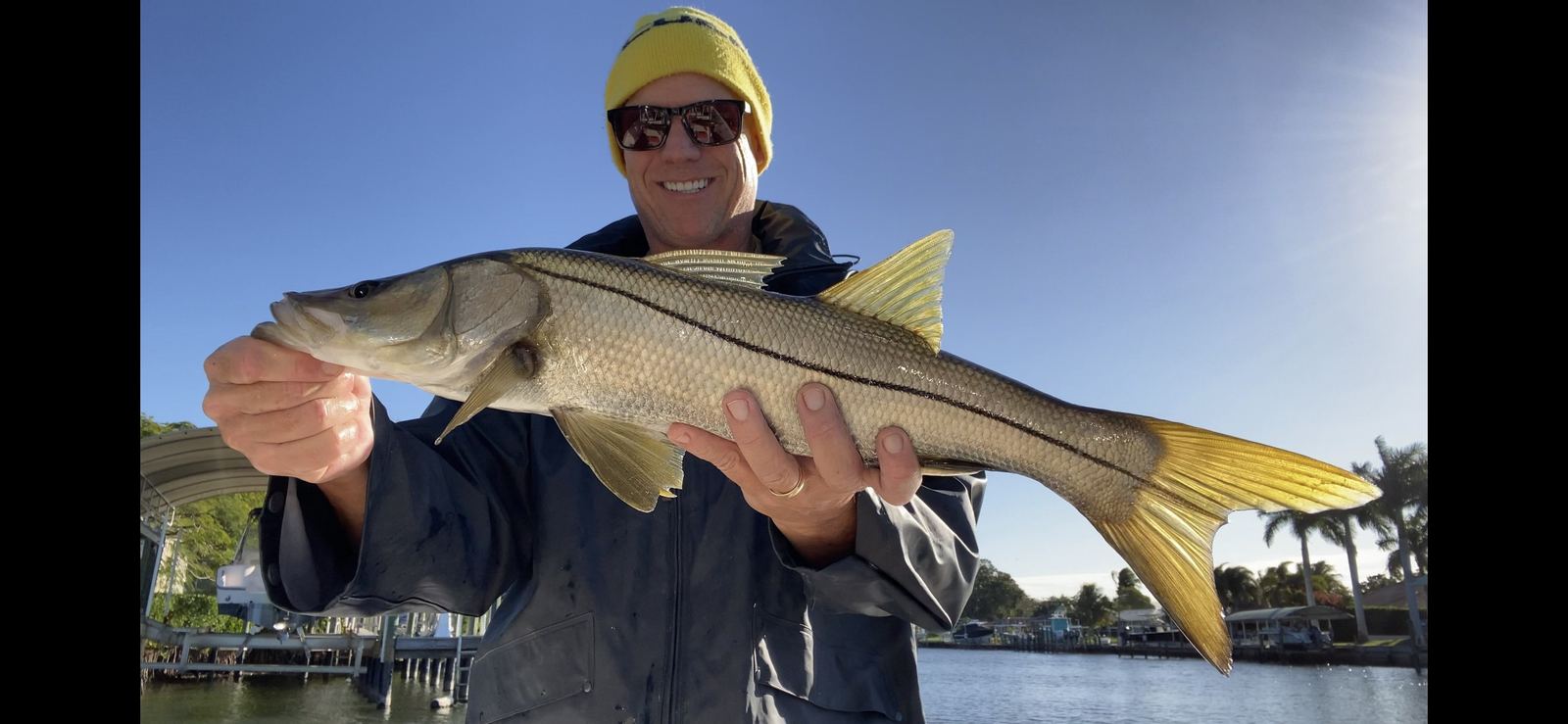 An angler in a knit cap holding a snook along a Sarasota canal shoreline