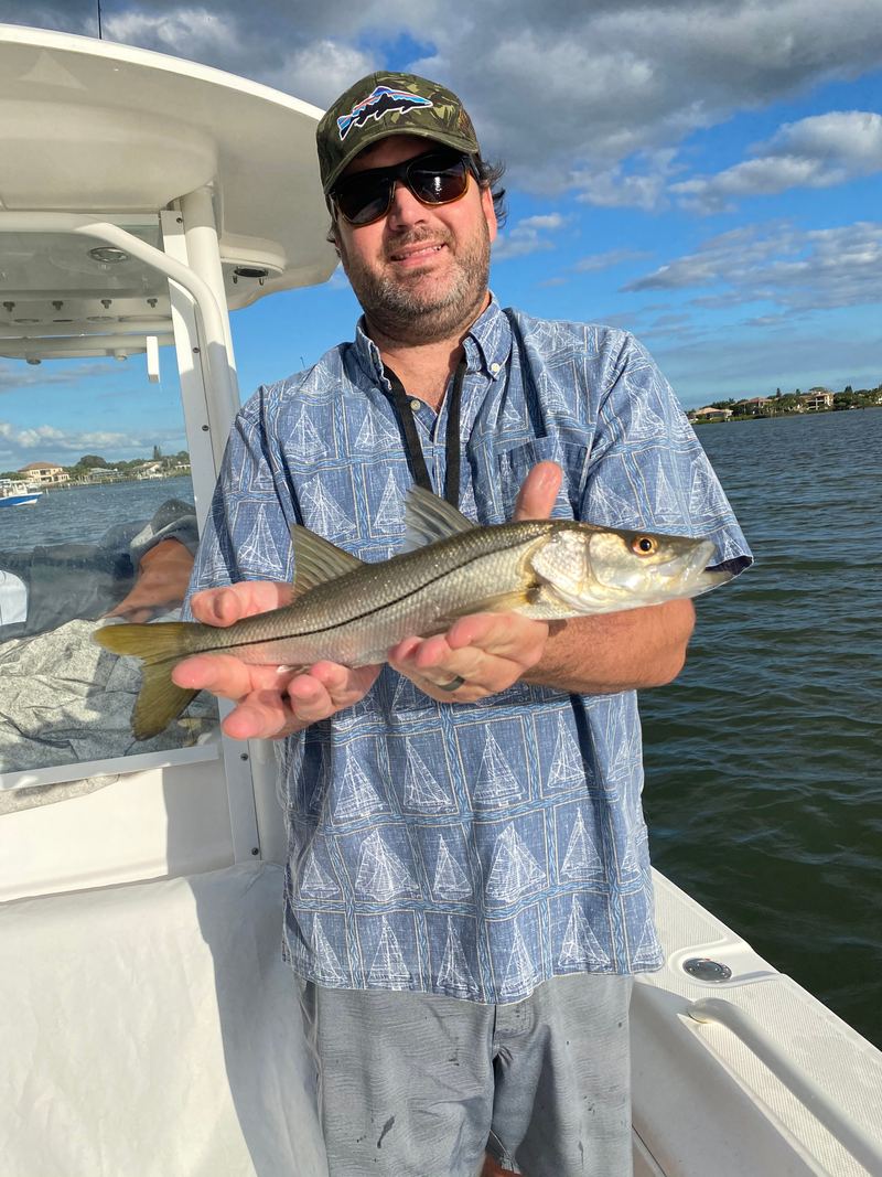 An angler in a short-sleeve shirt holding a small snook across both hands on a boat deck, bay and shoreline behind