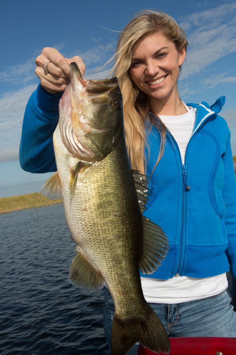 An angler holding a heavy largemouth bass upright by the jaw, a low marsh-grass shoreline and open water behind