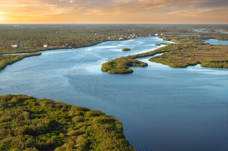 A wide tidal river winding through low green islands and marsh at last light, houses set back along the far bank